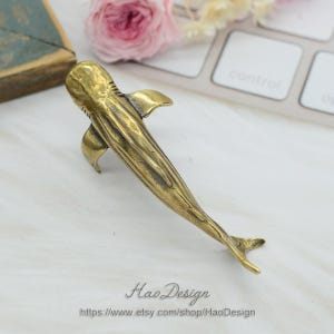 May include: A brass whale shark figurine with a detailed, textured surface. The figurine is positioned on a white surface.