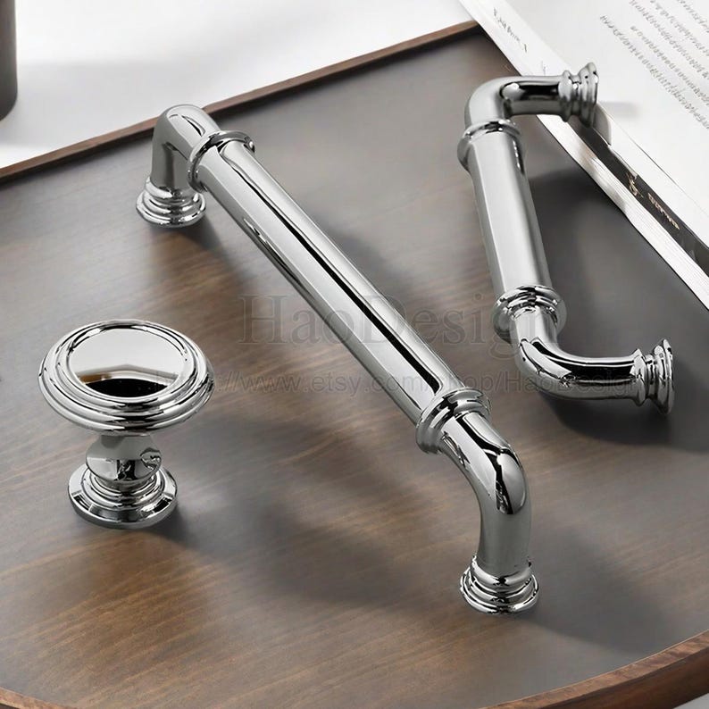 May include: A set of polished chrome cabinet hardware, including two bar pulls, a smaller pull, and a round knob. The hardware is displayed on a wooden surface, reflecting the bright light.