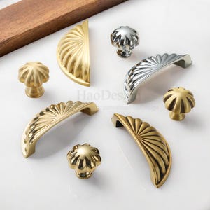 Może przedstawiać: Asortyment okuć do szafek w złotych i srebrnych wykończeniach. Kolekcja obejmuje r&oacute;żne style gałek i uchwyt&oacute;w, z rowkowanymi i wachlarzowymi wzorami. Okucia są prezentowane na białej powierzchni.