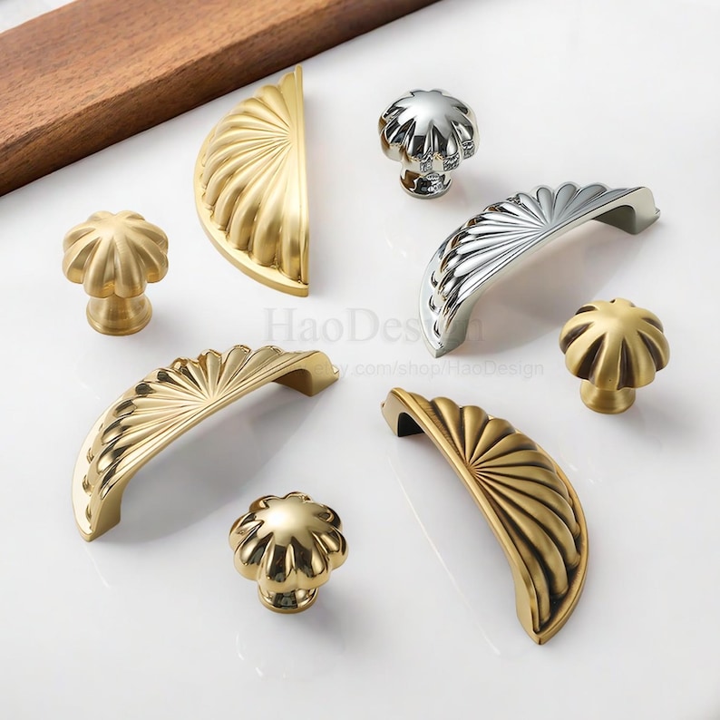 Może przedstawiać: Asortyment okuć do szafek w złotych i srebrnych wykończeniach. Kolekcja obejmuje r&oacute;żne style gałek i uchwyt&oacute;w, z rowkowanymi i wachlarzowymi wzorami. Okucia są prezentowane na białej powierzchni.