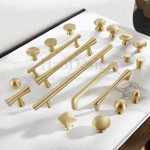 Puede incluir: Una colecci&oacute;n de herrajes dorados para armarios, que incluye pomos y tiradores, dispuestos sobre una superficie de m&aacute;rmol blanco y gris. Los herrajes presentan varios dise&ntilde;os, desde pomos redondos simples hasta tiradores de barra alargados, todos con acabado mate.