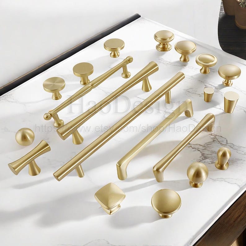 Puede incluir: Una colecci&oacute;n de herrajes dorados para armarios, que incluye pomos y tiradores, dispuestos sobre una superficie de m&aacute;rmol blanco y gris. Los herrajes presentan varios dise&ntilde;os, desde pomos redondos simples hasta tiradores de barra alargados, todos con acabado mate.