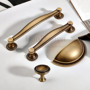 Puede incluir: Un juego de herrajes de latón antiguo para gabinetes, que incluye dos manijas curvas, un tirador en forma de media luna y un pequeño pomo. Los herrajes presentan un diseño de borde con cuentas detallado y un acabado cálido y envejecido.