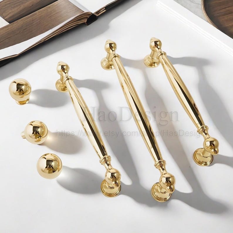 K&ouml;nnte beinhalten: Goldfarbenes Schrankbeschlags-Set, bestehend aus drei langen Griffen und drei runden Kn&ouml;pfen. Die Griffe haben ein schlankes, l&auml;ngliches Design mit dekorativen Enddetails. Die Kn&ouml;pfe sind kugelf&ouml;rmig. Das Set wird auf einer wei&szlig;en Oberfl&auml;che pr&auml;sentiert.