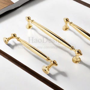 K&ouml;nnte beinhalten: Vier goldfarbene Schrankgriffe mit klassischem Design. Die Griffe haben eine glatte, polierte Oberfl&auml;che und sind auf einer wei&szlig;en Oberfl&auml;che angeordnet. Der Text "HaoDesign" und eine Website-Adresse sind sichtbar.