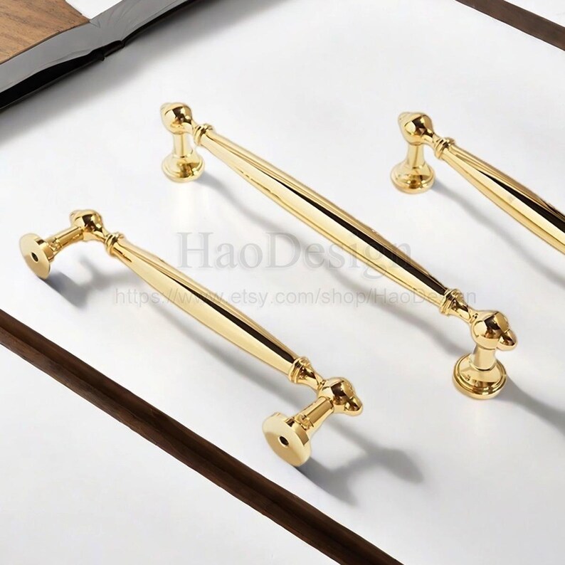 K&ouml;nnte beinhalten: Vier goldfarbene Schrankgriffe mit klassischem Design. Die Griffe haben eine glatte, polierte Oberfl&auml;che und sind auf einer wei&szlig;en Oberfl&auml;che angeordnet. Der Text "HaoDesign" und eine Website-Adresse sind sichtbar.