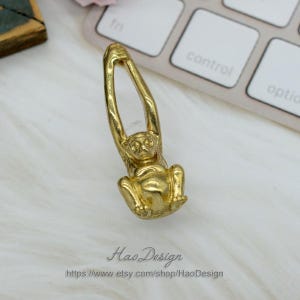 K&ouml;nnte beinhalten: Ein goldfarbener Affenanh&auml;nger, der an einer Schlaufe h&auml;ngt. Der Affe ist in einer h&auml;ngenden Position dargestellt, mit den Armen &uuml;ber dem Kopf erhoben.