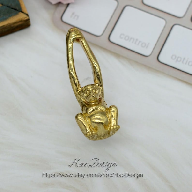 K&ouml;nnte beinhalten: Ein goldfarbener Affenanh&auml;nger, der an einer Schlaufe h&auml;ngt. Der Affe ist in einer h&auml;ngenden Position dargestellt, mit den Armen &uuml;ber dem Kopf erhoben.