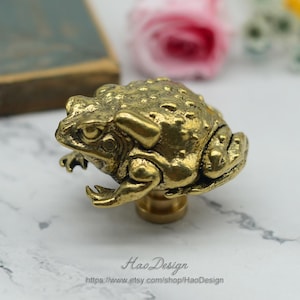 May include: A gold-toned frog-shaped cabinet knob or drawer pull. The frog is detailed with textured skin and has a realistic appearance.