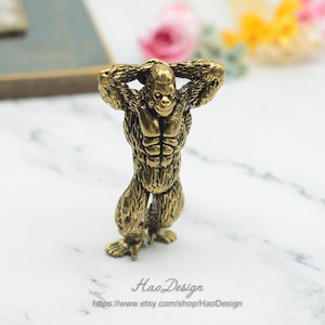 May include: A gold-coloured metal figurine of a gorilla standing with its arms raised above its head. The gorilla has a detailed, muscular body and a textured surface.