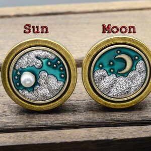 Puede incluir: Dos pendientes redondos de latón antiguo. Uno presenta un diseño de sol con nubes y la palabra "Sun". El otro tiene un diseño de luna y estrellas con nubes y la palabra "Moon". Ambos tienen un fondo turquesa.