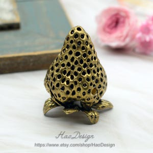 Op de afbeelding: Een gouden metalen ringhouder in de vorm van een aardbei. De aardbei is gedetailleerd met een textuur en kleine gaatjes.