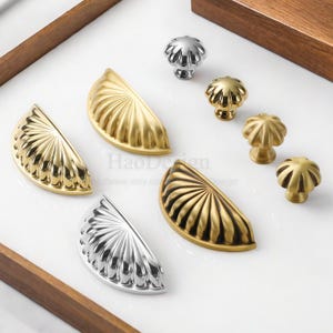 May include: A selection of decorative cabinet hardware, featuring gold and silver half-moon pulls and mushroom-shaped knobs. The hardware has a fluted design and is displayed on a white surface.