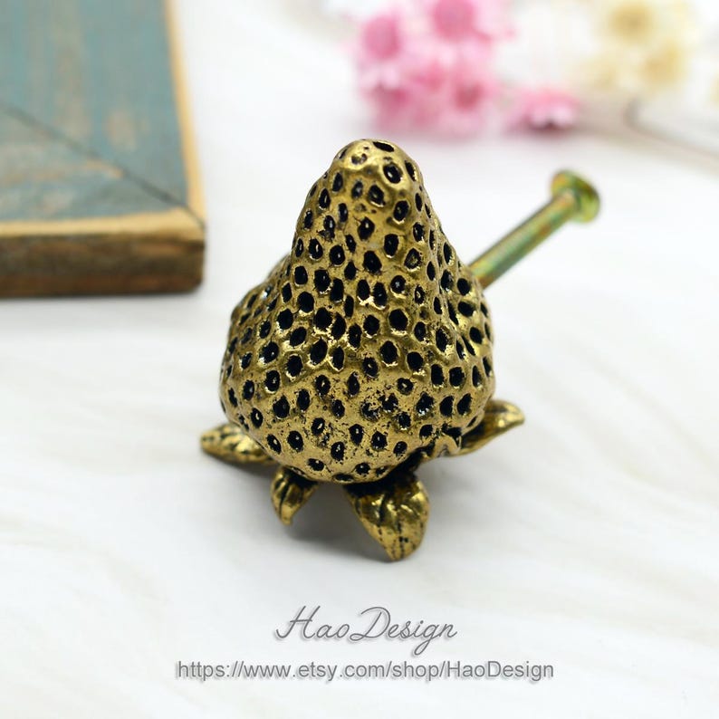 Op de afbeelding: Gouden metalen kastknop in de vorm van een aardbei met een getextureerd oppervlak en een schroef voor installatie.