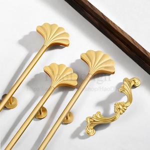 May include: Gold-colored cabinet hardware featuring a fan-shaped design. The image shows several long pulls and a shorter handle with ornate detailing. The hardware is set against a white background with a dark wood accent.