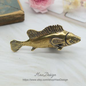 Könnte beinhalten: Eine goldfarbene Fischfigur aus Metall mit detaillierten Schuppen und Flossen. Der Fisch blickt nach rechts und hat einen leicht geöffneten Mund.