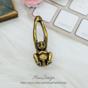 K&ouml;nnte beinhalten: Eine goldfarbene Metallfigur eines Affen, der an einem Ast h&auml;ngt. Der Affe sitzt mit erhobenen Armen &uuml;ber dem Kopf.