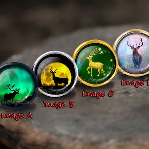Puede incluir: Cuatro cabujones redondos de vidrio con engastes de metal dorado, plateado y negro. Cada cabujón presenta una imagen diferente de un ciervo en un entorno de bosque. El primer cabujón muestra un ciervo silueteado contra una aurora boreal verde y azul. El segundo cabujón muestra un ciervo silueteado contra una luna llena. El tercer cabujón muestra un ciervo dorado con cuernos brillantes en un bosque verde. El cuarto cabujón muestra un ciervo marrón con cuernos brillantes en una galaxia rosa y azul.