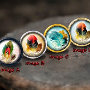 Könnte beinhalten: Vier runde Glascabochons mit goldenen und silbernen Fassungen, jede mit einer anderen Aquarellmalerei einer bunten Feder.