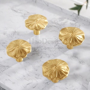 May include: Four gold-colored cabinet knobs with a lotus flower design. The knobs are made of metal and have a detailed, textured surface. The knobs are arranged on a white and gray marble-patterned surface.