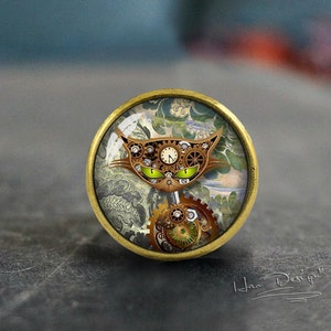 Könnte beinhalten: Ein runder, messingfarbener, dekorativer Knopf mit einem Glaseinsatz, der eine Katze im Steampunk-Stil mit Zahnrädern und einem Zifferblatt zeigt. Die Katze hat grüne Augen und ist vor einem Hintergrund aus wirbelnden Mustern platziert.