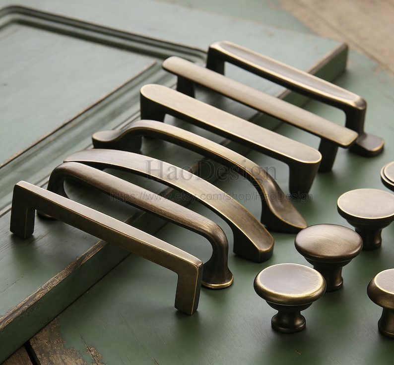 May include: A selection of antique brass cabinet hardware, featuring pull handles and round knobs. The handles are in various shapes, some straight and some curved. The hardware is displayed on a green surface.