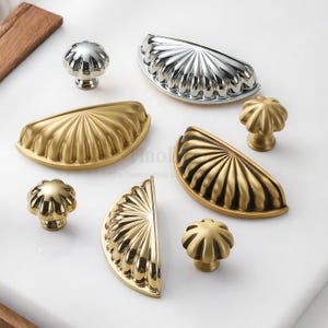 Może przedstawiać: Kolekcja dekoracyjnych okuć do szafek, w tym uchwyty w kształcie muszli i okrągłe gałki. Okucia są dostępne w złotym i srebrnym wykończeniu. Uchwyty mają wachlarzowaty kształt, a gałki wyglądają jak kwiaty.