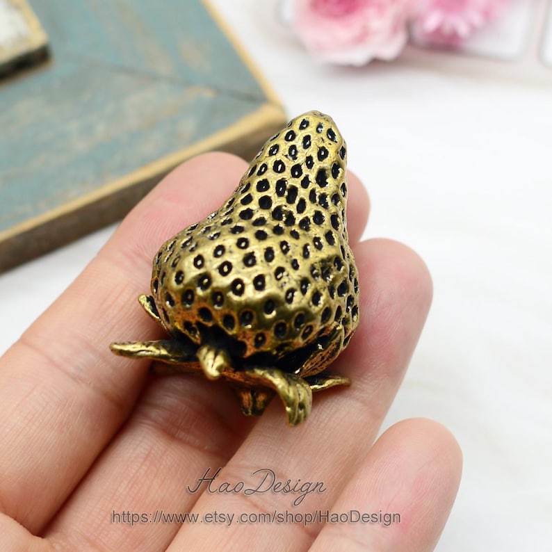 Op de afbeelding: Een gouden metalen aardbeienfiguur met een gedetailleerd, gestructureerd oppervlak. De aardbei ligt op een hand.