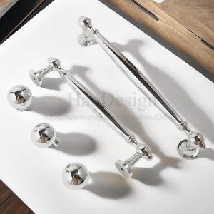 K&ouml;nnte beinhalten: Gl&auml;nzendes silbernes Schrankbeschlagsset. Enth&auml;lt zwei lange Zuggriffe und drei runde Kn&ouml;pfe. Die Griffe haben ein geschwungenes Design mit dekorativen Enden. Die Kn&ouml;pfe sind kugelf&ouml;rmig. Das Set befindet sich auf einer wei&szlig;en Oberfl&auml;che.