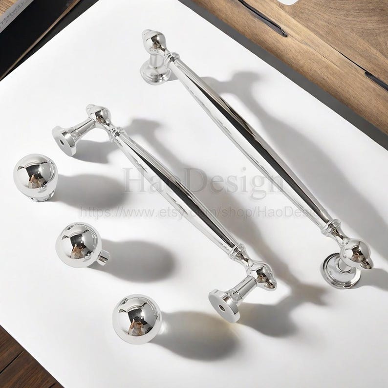 K&ouml;nnte beinhalten: Gl&auml;nzendes silbernes Schrankbeschlagsset. Enth&auml;lt zwei lange Zuggriffe und drei runde Kn&ouml;pfe. Die Griffe haben ein geschwungenes Design mit dekorativen Enden. Die Kn&ouml;pfe sind kugelf&ouml;rmig. Das Set befindet sich auf einer wei&szlig;en Oberfl&auml;che.