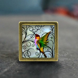 Può includere: Un colibrì colorato con piume verdi, rosa e gialle è raffigurato in un cammeo di vetro con un design di viticci a spirale in bianco e nero. Il cammeo è incastonato in una cornice di metallo dorato.