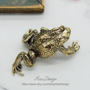 May include: A gold-colored metal frog figurine with a detailed textured surface. The frog is posed in a sitting position with its legs outstretched.