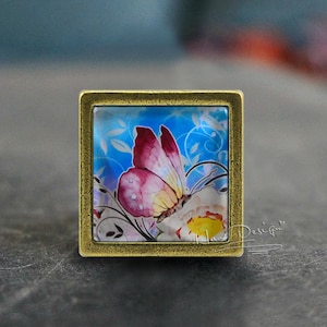 Puede incluir: Un marco cuadrado de metal dorado con un inserto de vidrio que presenta una pintura de acuarela de una mariposa rosa y amarilla sobre un fondo azul con acentos florales blancos.