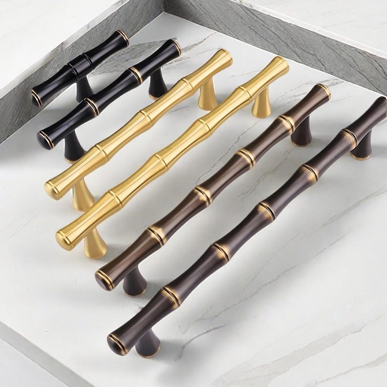 May include: A collection of bamboo-style cabinet pulls in various finishes: black, gold, and bronze. The pulls are arranged on a white marble surface, showcasing their decorative design and potential for home decor projects.