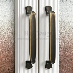 May include: Two bronze-coloured door handles with a vertical, elongated design. The handles are mounted on a white door with vertical grooves. The background features a textured glass panel. The Etsy shop name and URL are visible.