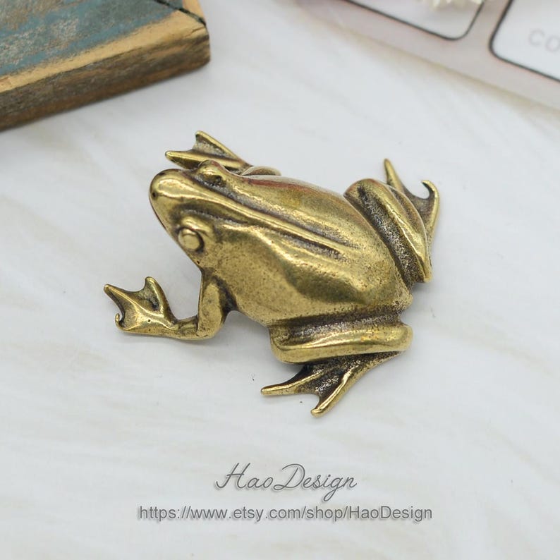 Może przedstawiać: Mała, solidna figurka żaby z mosiądzu. Żaba siedzi z rozłożonymi nogami i lekko pochyloną głową. Figurka jest złotego koloru i ma gładkie, polerowane wykończenie.