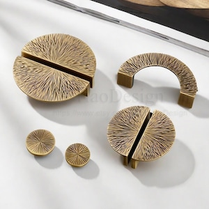 May include: A collection of brass cabinet hardware, including two half-circle handles, a curved handle, and two round knobs. The hardware features a textured, sunburst-like pattern. The pieces are displayed on a white surface.
