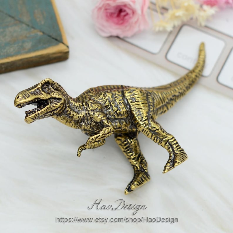 Pode incluir: Uma figurinha de dinossauro de metal dourado. O dinossauro est&aacute; em p&eacute; sobre suas quatro patas com a cabe&ccedil;a ligeiramente inclinada para o lado. A figurinha &eacute; detalhada com escamas texturizadas e uma superf&iacute;cie texturizada.