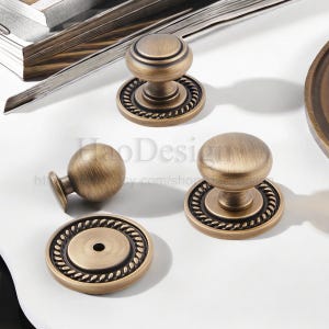 May include: Antique brass cabinet knobs and pulls. The knobs have a round shape with a decorative rope-like border. The hardware is made of metal and has a vintage aesthetic. The knobs are shown on a white surface.