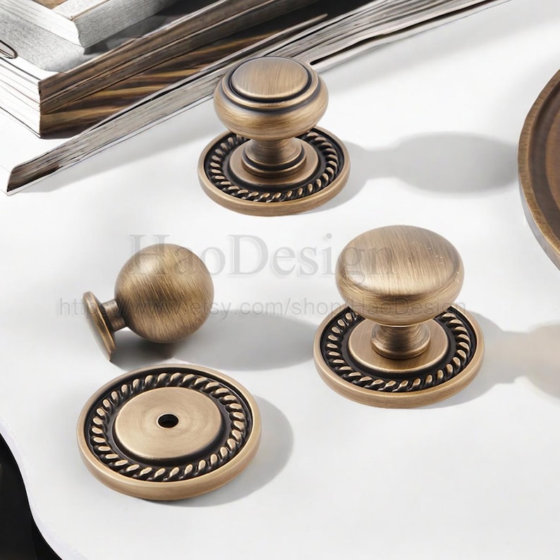 May include: Antique brass cabinet knobs and pulls. The knobs have a round shape with a decorative rope-like border. The hardware is made of metal and has a vintage aesthetic. The knobs are shown on a white surface.