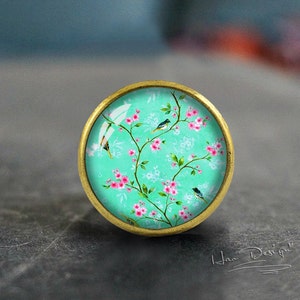 Puede incluir: Un pomo redondo de color dorado con un fondo azul turquesa y un patrón floral. El patrón presenta flores rosas y pequeños pájaros negros en ramas.