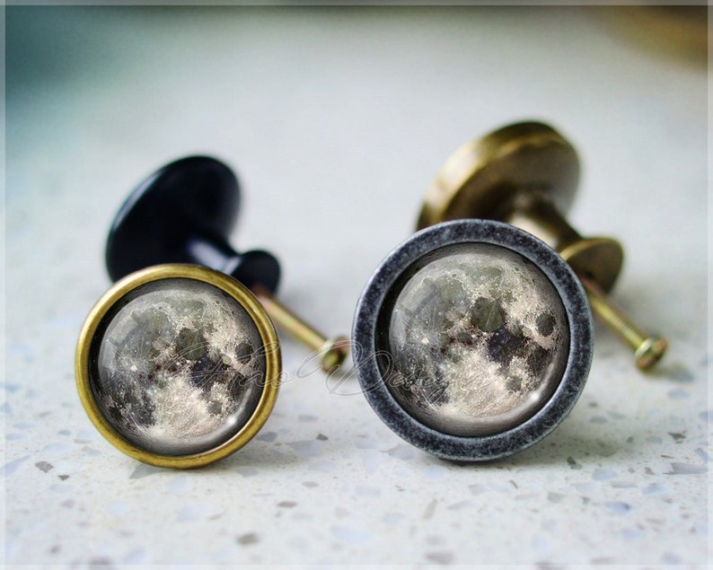 May include: Two cabinet knobs with a moon image. The knobs are made of metal with a black and gray finish. The moon image is in shades of gray and white.