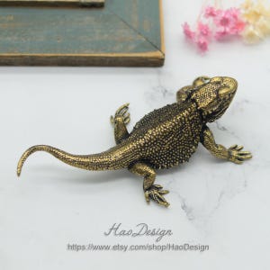 May include: A gold-colored metal figurine of a bearded dragon lizard. The figurine is detailed with textured scales and has a curled tail.