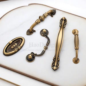 以下が含まれることがあります： アンティーク真鍮製のキャビネット用プルとハンドルの詰め合わせ。コレクションには、楕円形、曲線、バータイプのデザインがあり、装飾的なディテールが施されています。ハードウェアは白い表面に表示され、さまざまなスタイルと仕上げが示されています。