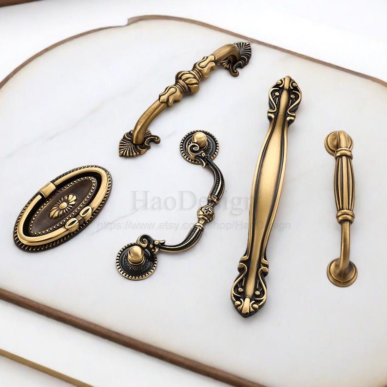 以下が含まれることがあります： アンティーク真鍮製のキャビネット用プルとハンドルの詰め合わせ。コレクションには、楕円形、曲線、バータイプのデザインがあり、装飾的なディテールが施されています。ハードウェアは白い表面に表示され、さまざまなスタイルと仕上げが示されています。