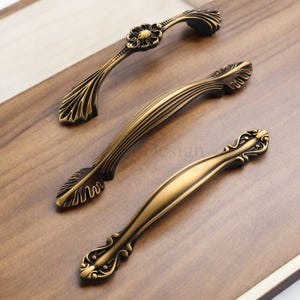 以下が含まれることがあります： 装飾的な花と葉のデザインが施された、3つのアンティークブロンズのキャビネットプル。プルはさまざまなサイズと形状で、暖かく、古びた仕上がりです。ハードウェアは、明るい茶色の木製の表面に表示されます。