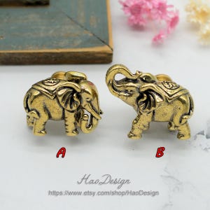 Puede incluir: Dos pomos de armario en forma de elefante de color dorado con detalles intrincados. Los pomos están sobre una superficie blanca con un fondo gris claro.