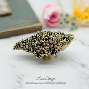 Könnte beinhalten: Ein goldfarbener Schrank-Knopf in Form eines Alligators mit schwarzen Augen. Der Knopf ist mit strukturierten Schuppen verziert und hat ein leichtes Lächeln.