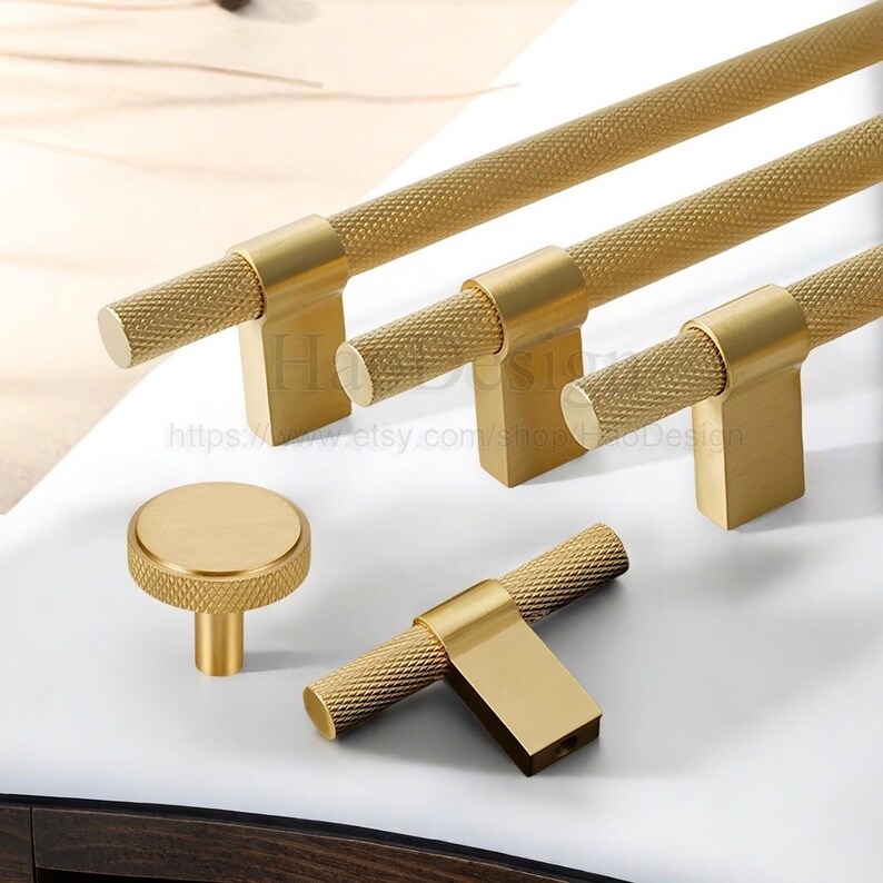 May include: A collection of gold-toned cabinet hardware, including bar pulls and a round knob. The pulls have a textured, diamond-patterned grip and rectangular mounting plates. The knob also features the diamond pattern. The hardware is displayed on a white surface.