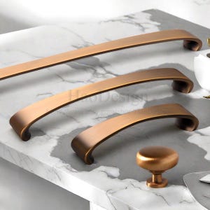 以下が含まれることがあります： ブロンズ色のキャビネット金具セット。3つのカーブしたハンドルと丸いノブが含まれています。白とグレーの大理石の表面に表示されています。ハンドルはサイズが異なり、統一感のあるノブが付いています。